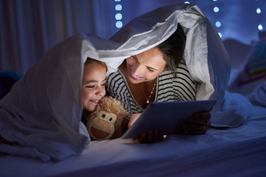 mother-and-young-daughter-under-blanket-at-night-using-bright-tablet