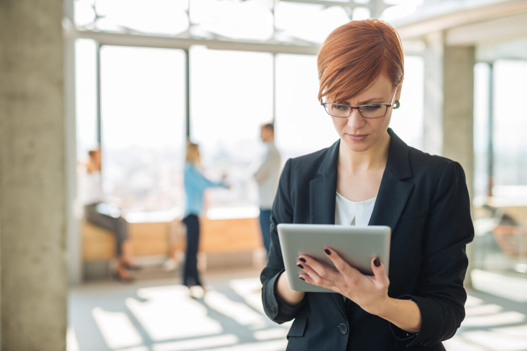 businesswoman-wearing-blazer-and-glasses-looking-at-tablet-while-standing-in-office-with-coworkers-in-background