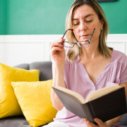 Middle aged woman putting on glasses to read a book.