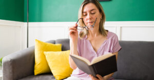 Middle aged woman putting on glasses to read a book.