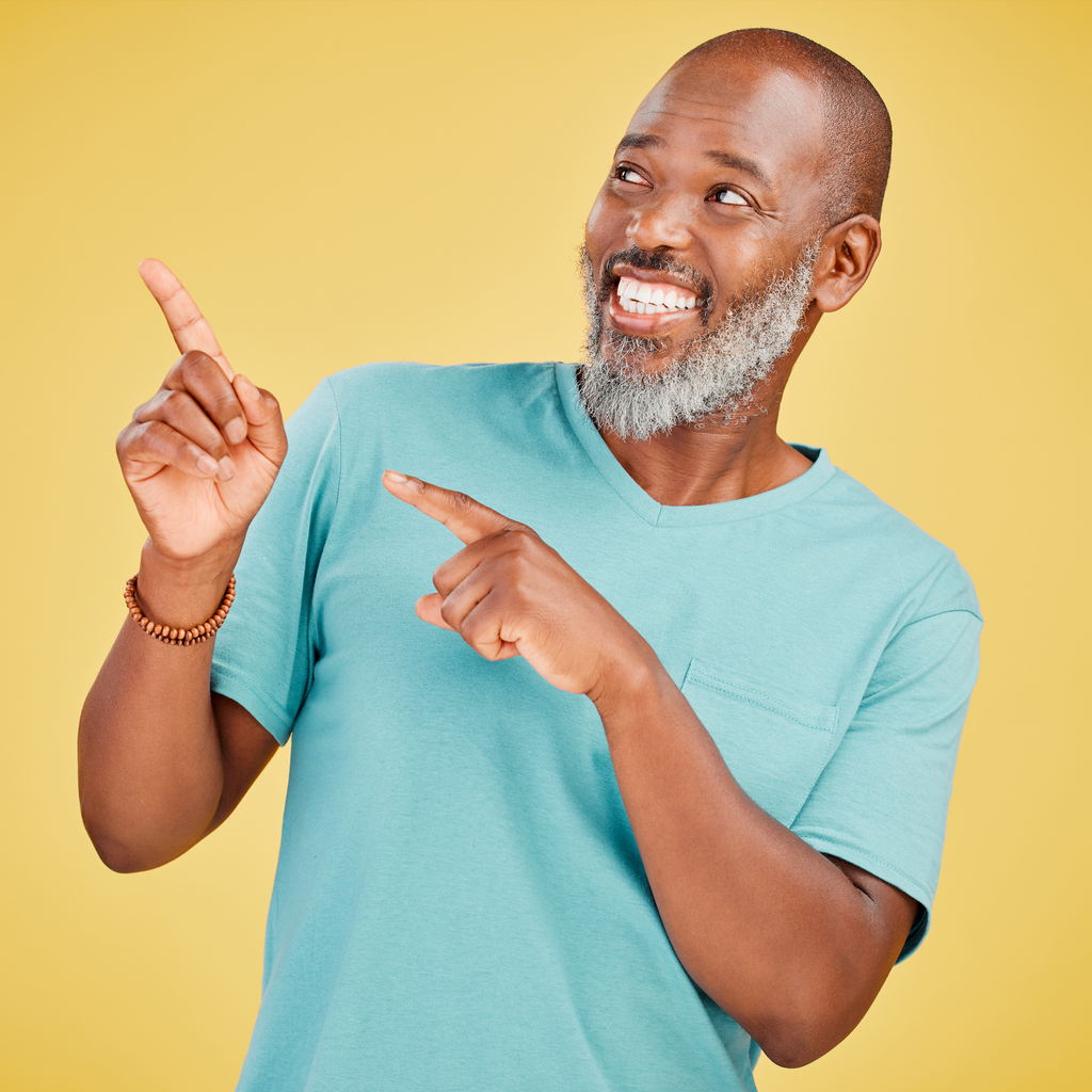 Mature african man smiling and pointing in a direction against a yellow studio background. Black guy looking happy and making a pointing gesture reacting with a smile while looking cheerful and happy. 